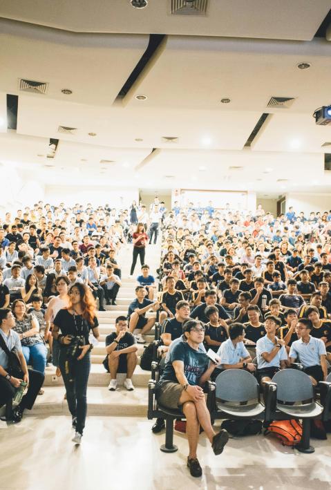 Frau in schwarzem Kleid mit Rundhalsausschnitt geht in der N&auml;he eines Stuhls in einem gro&szlig;en Konferenzraum mit vielen anderen Menschen 