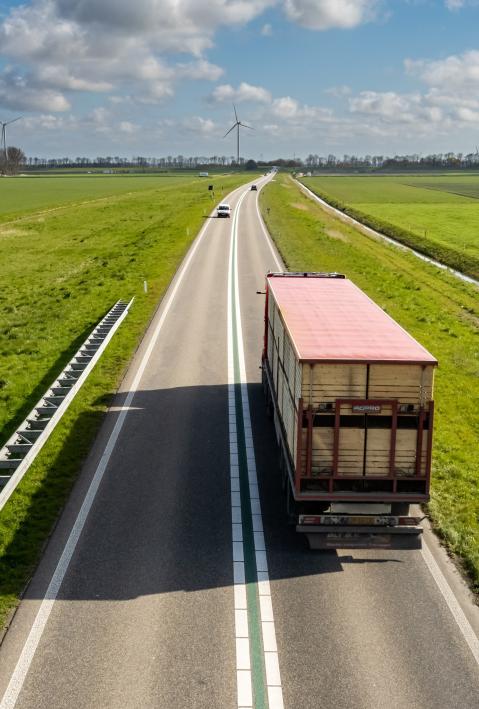 A truck drives between green meadows towards a hilltop with wind turbines. 