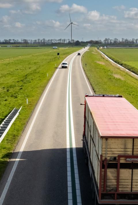 Ein Lastwagen f&auml;hrt zwischen gr&uuml;nen Wiesen auf eine Bergkuppe mit Windr&auml;dern zu. 