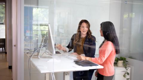 Zwei Frauen am Stechtisch in einer Arbeitssituation vor Laptop. 