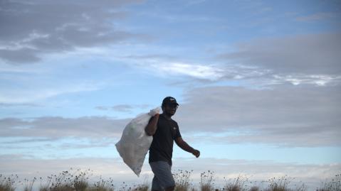 a man in the dunes near a beach carrying a big bag full of trash with the horizon in the background